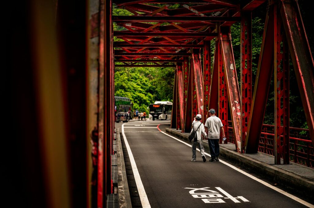 Photo of two people walking across a red iron bridge in Taiwan with greenery and a bus in the background.